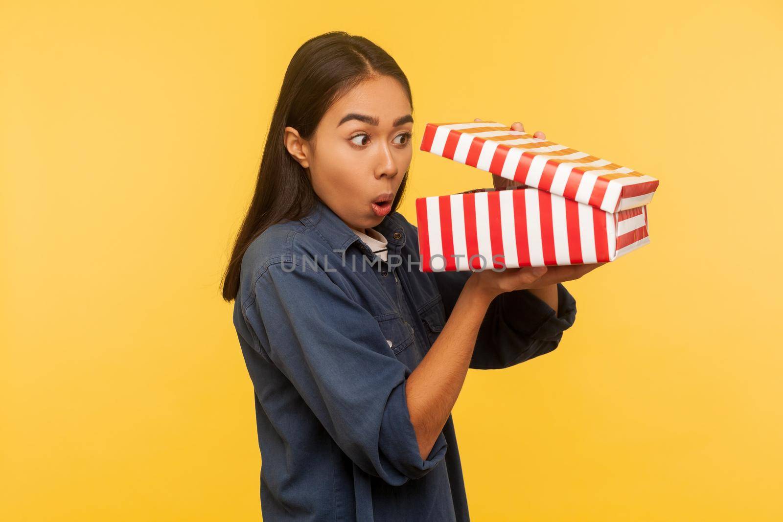 Portrait of amazed funny girl in denim shirt opening gift and looking into box with shocked astonished expression, found unbelievable present surprise. indoor studio shot isolated on yellow background