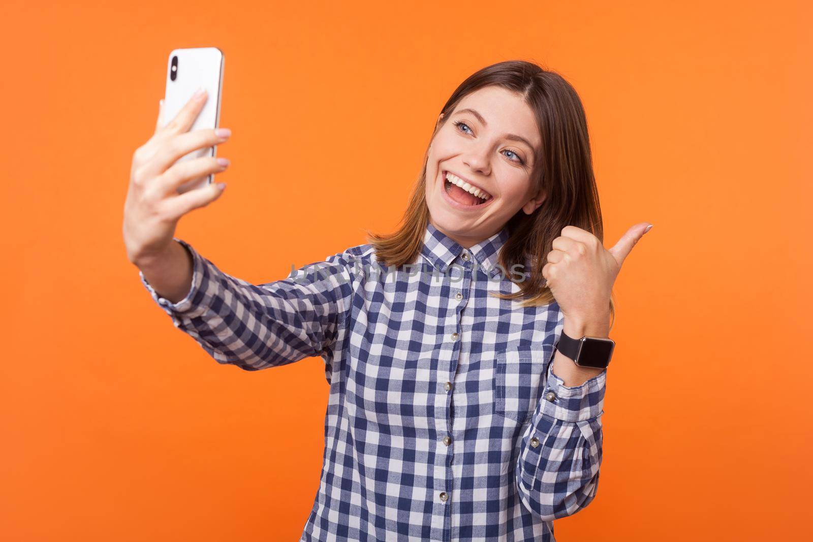 Thumbs up. Portrait of lovely brunette woman with charming smile wearing checkered shirt using cellphone, showing like gesture while making video call. indoor studio shot isolated on orange background