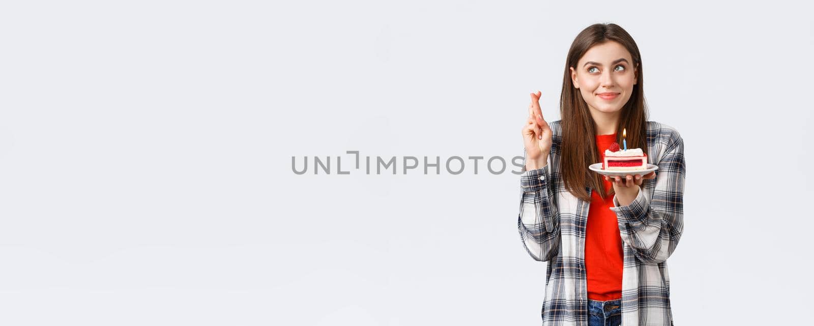 People lifestyle, holidays and celebration, emotions concept. Dreamy hopeful birthday girl in casual clothes, cross fingers and look up making wish, holding b-day cake, blowing candle.