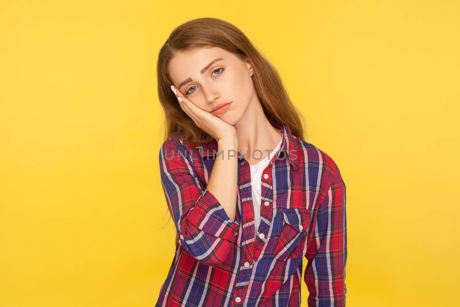 I'm bored. Portrait of depressed ginger girl in checkered shirt leaning on cheek and looking with indifferent dull expression, tired of boring story. indoor studio shot isolated on yellow background