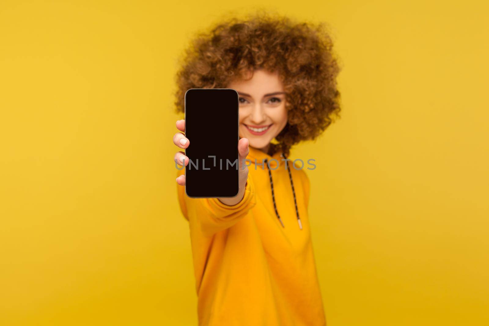 Portrait of cheerful curly-haired woman with engaging smile showing cellphone with empty display, mock up for app advertise, mobile web service. indoor studio shot isolated on yellow background