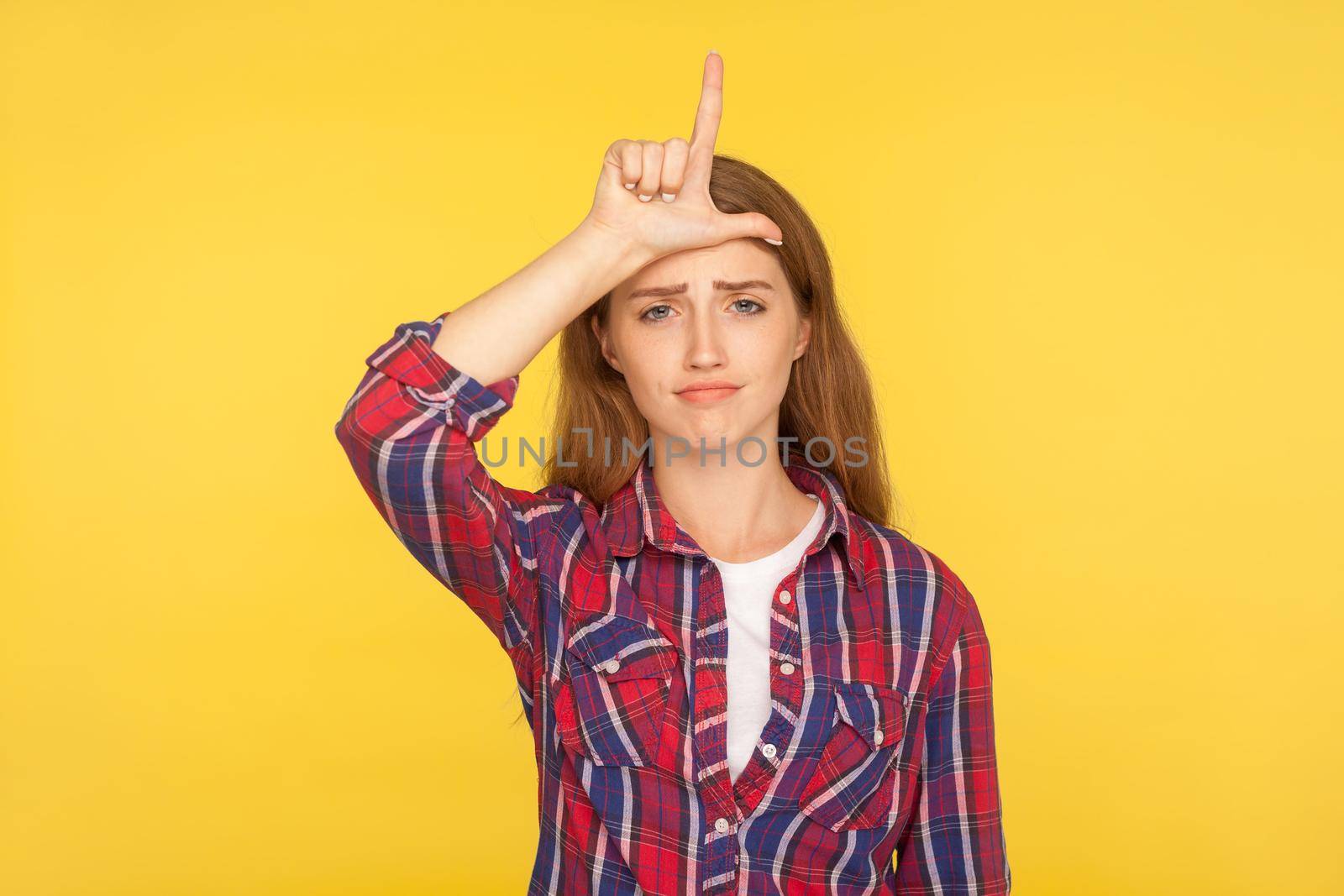 Portrait of frustrated unlucky ginger girl in casual shirt showing loser gesture, L finger sign on forehead and looking with sarcastic gloomy expression. studio shot isolated on yellow background