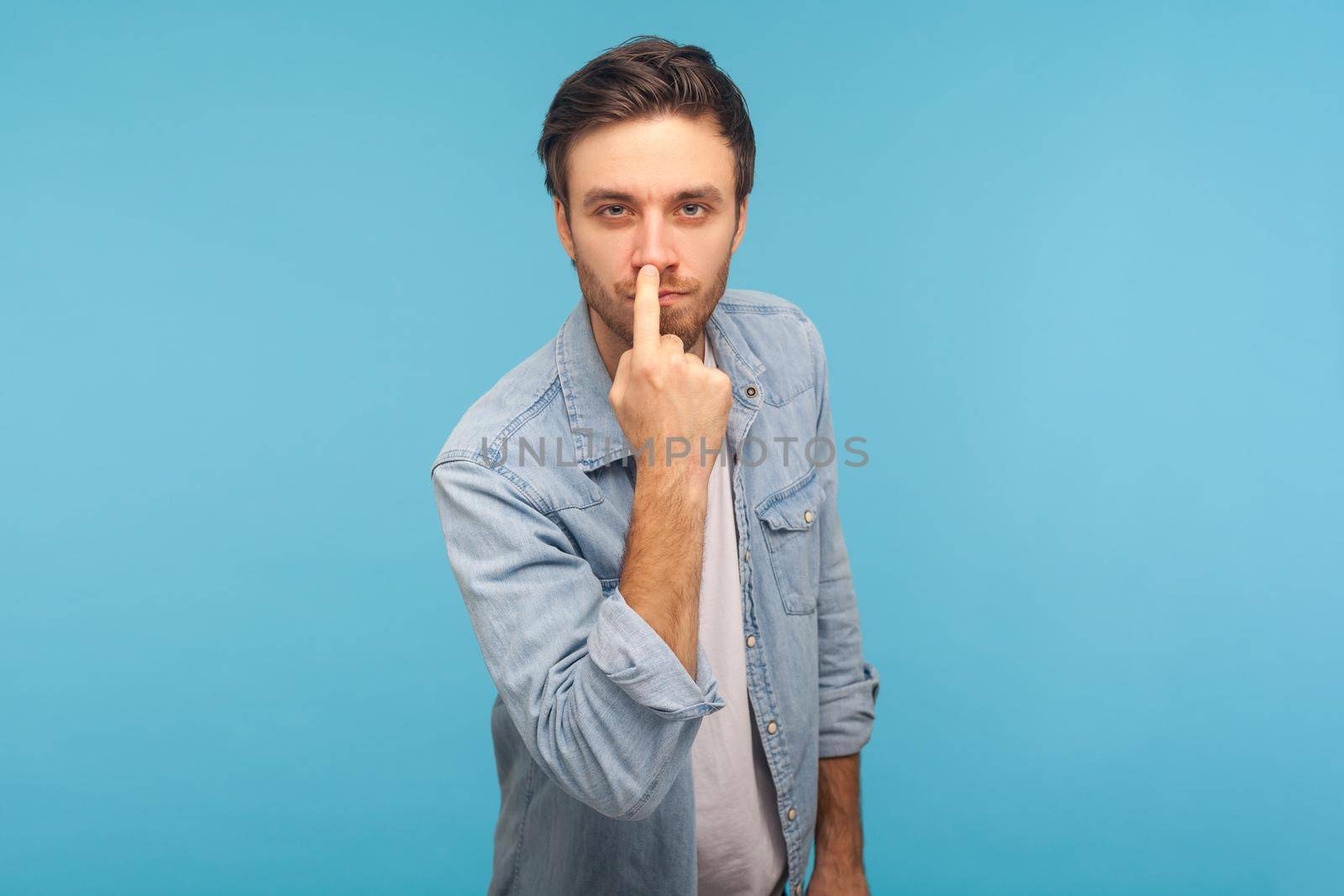 Don't lie to me. Portrait of man in worker denim shirt touching nose, showing liar gesture, angry about falsehood, outright deception, fake news. indoor studio shot isolated on blue background