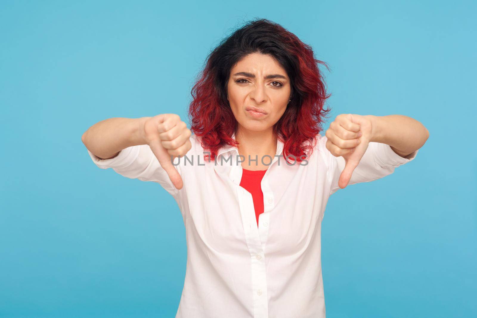 Dislike, bad review. Portrait of frustrated dissatisfied hipster woman with fancy red hair showing thumbs down, expressing disapproval, critique. indoor studio shot isolated on blue background