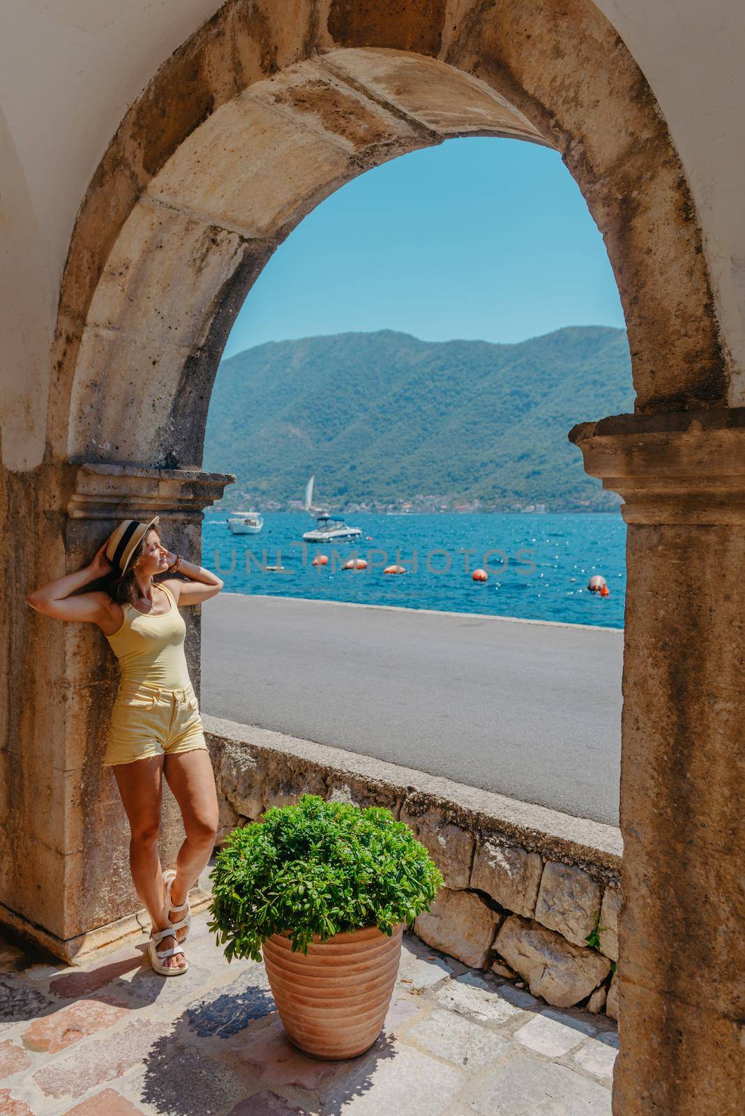 Summer photo shoot on the streets of Kotor, Montenegro. Beautiful girl in white dress and hat. smiling tourist girl with hat. Spectacular view of Montenegro with copy space. fashion outdoor photo of beautiful sensual woman with blond hair in elegant dress and straw hat and bag, posing in Montenego's city Perast by Andrii_Ko