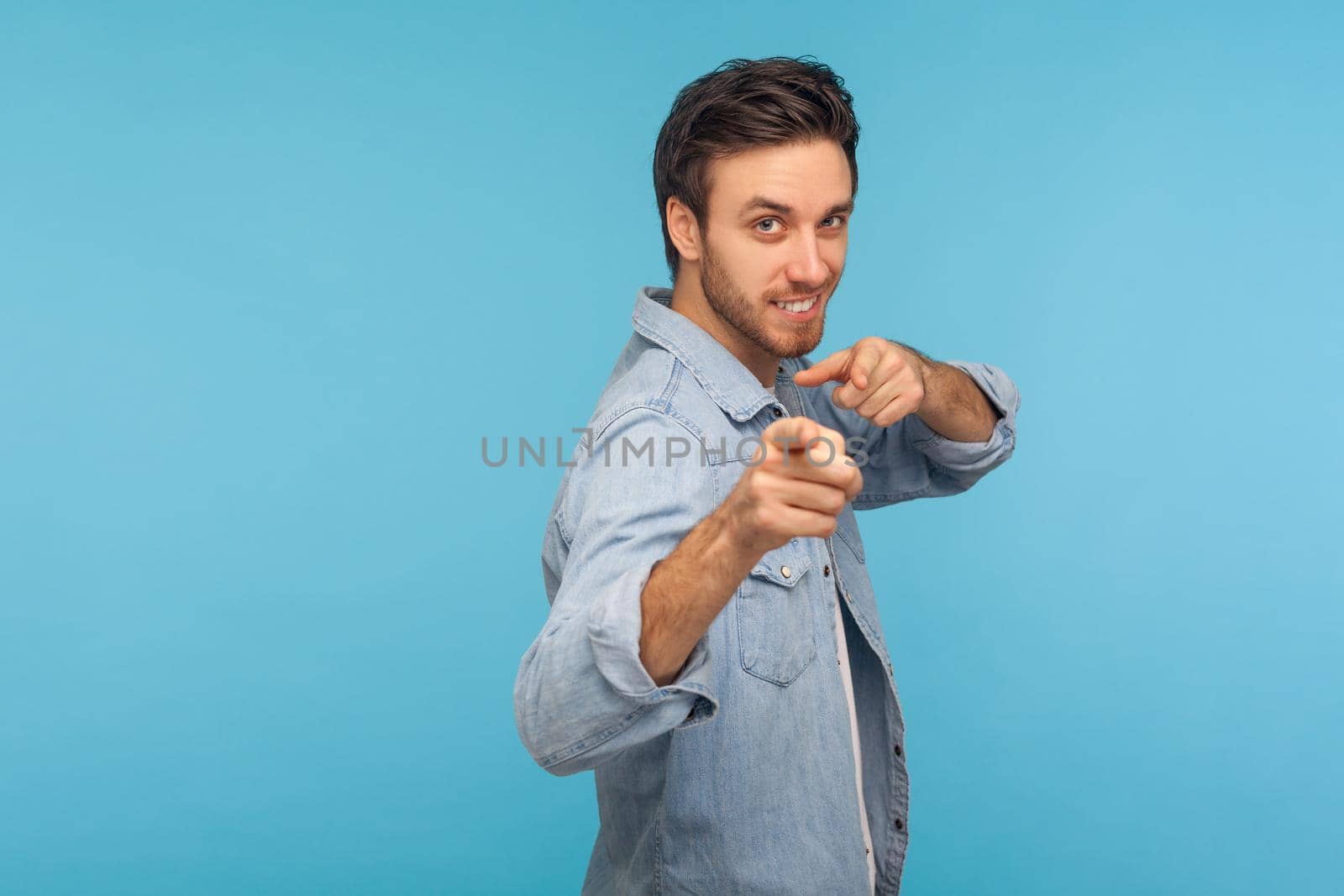 We need you. Portrait of cheerful happy man in worker denim shirt noticing and pointing finger to camera, choosing lucky winner, smiling joyfully. indoor studio shot isolated on blue background
