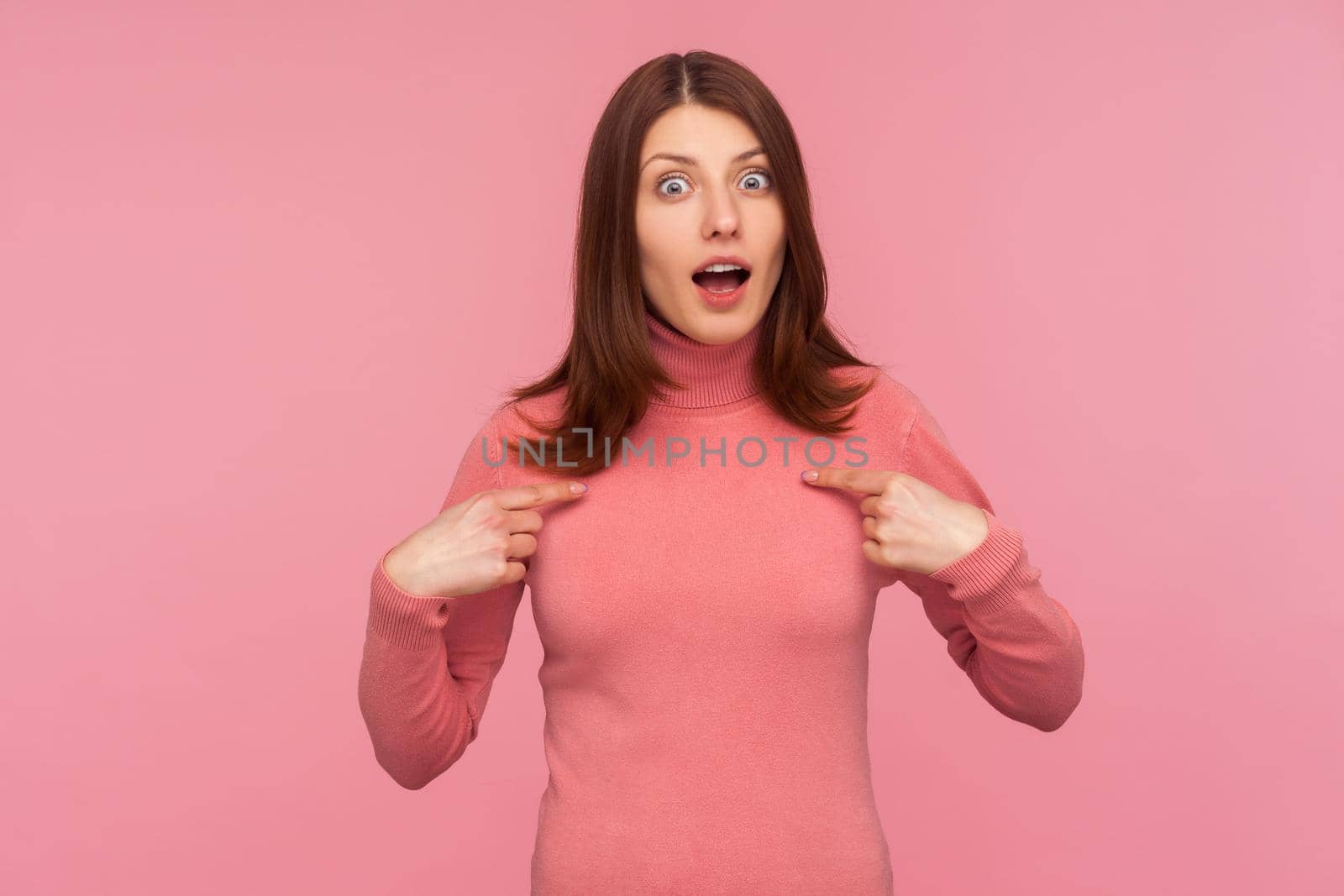 Insecure shocked woman with brown hair in pink sweater pointing fingers on herself, surprised with her participation, cant believe in sudden victory. Indoor studio shot isolated on pink background