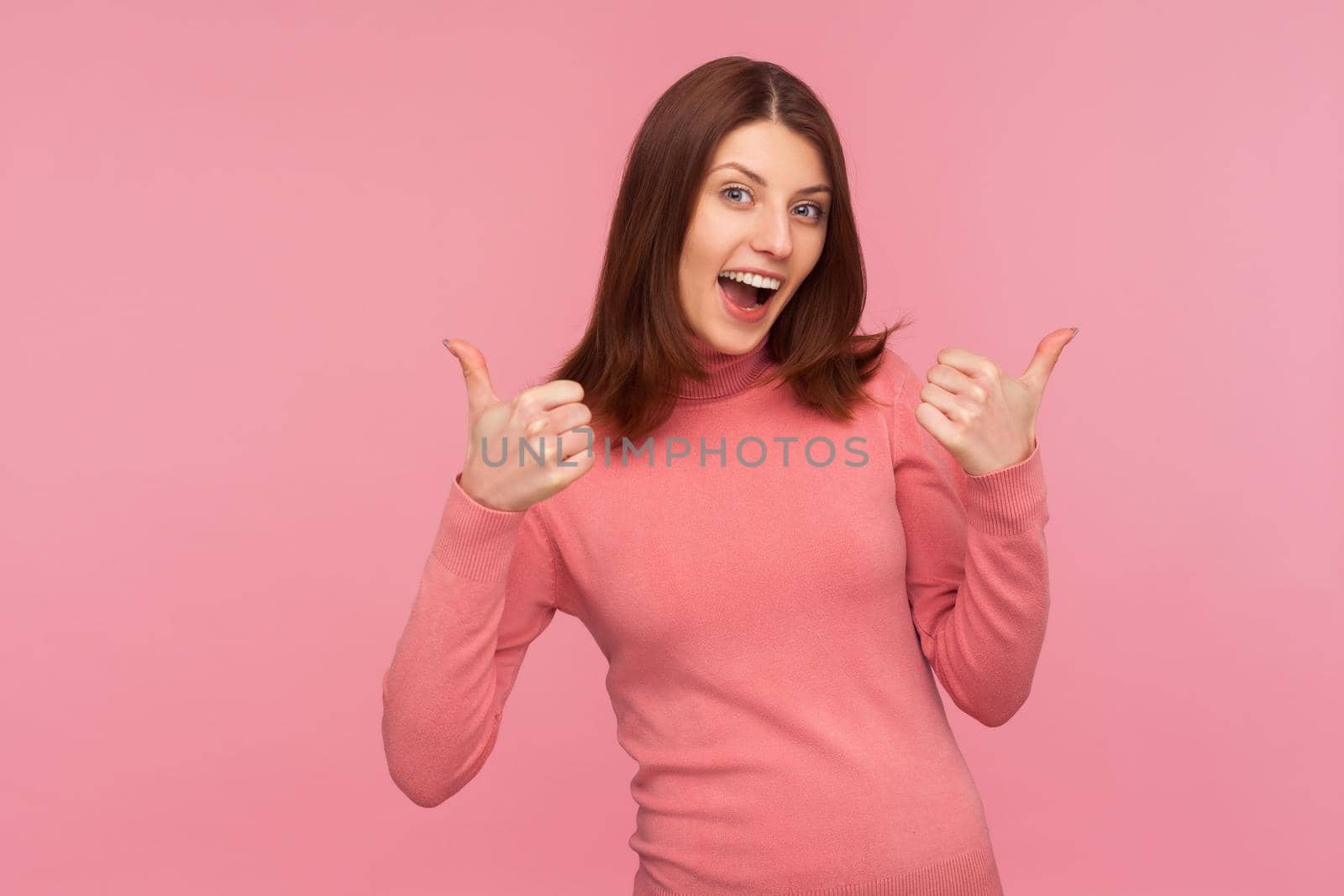 Well done. Extremely happy excited brunette woman showing thumbs up, like gesture, looking at camera with satisfied expression. Indoor studio shot isolated on pink background