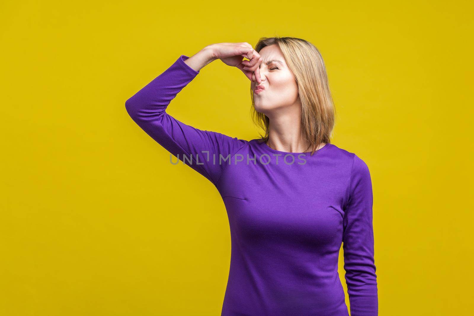 Bad smell. Portrait of young woman in purple dress standing pinching her nose with fingers to hold breath, disgusted by stinky intolerable smell. indoor studio shot isolated on yellow background