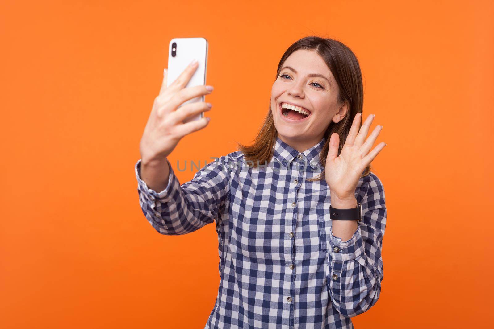 Hello. Portrait of adorable brunette woman with charming smile wearing checkered shirt using cellphone, showing Hi gesture while making video call. indoor studio shot isolated on orange background