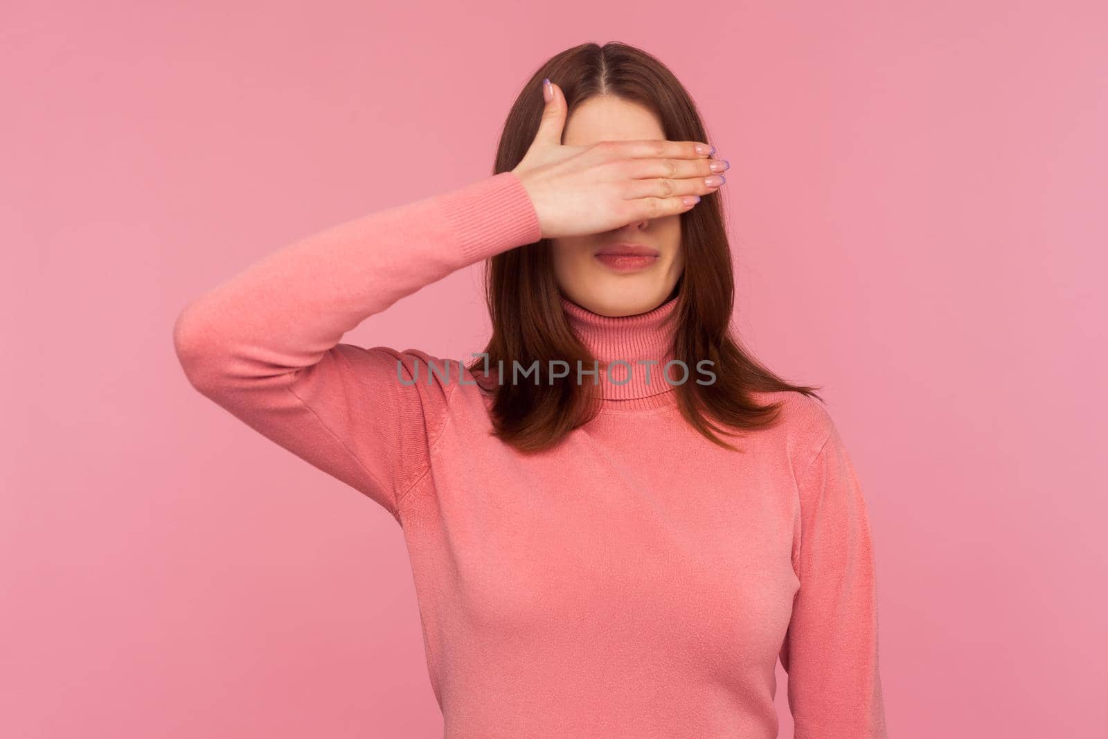 Ashamed confused woman with brown hair closing eyes with hand, hiding, ignoring dont want to see. Indoor studio shot isolated on pink background