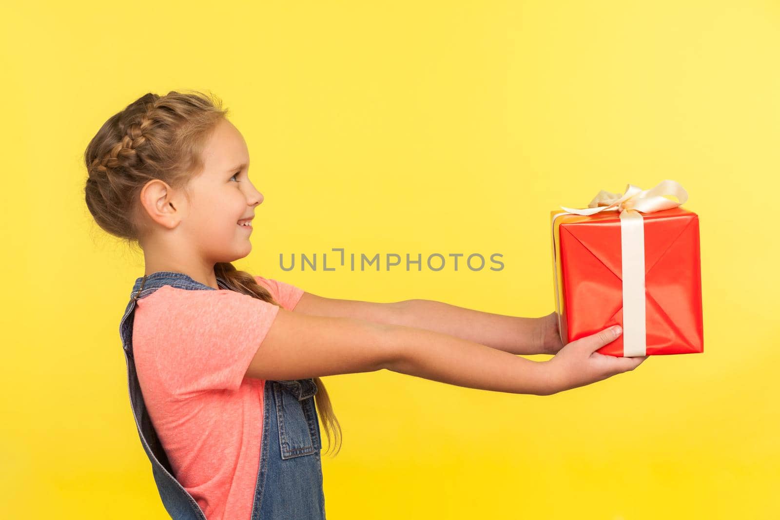 Take it. Side view of cute little girl with braid looking up and giving decorated present, generous child sharing christmas gift, kindness and charity. indoor studio shot isolated on yellow background