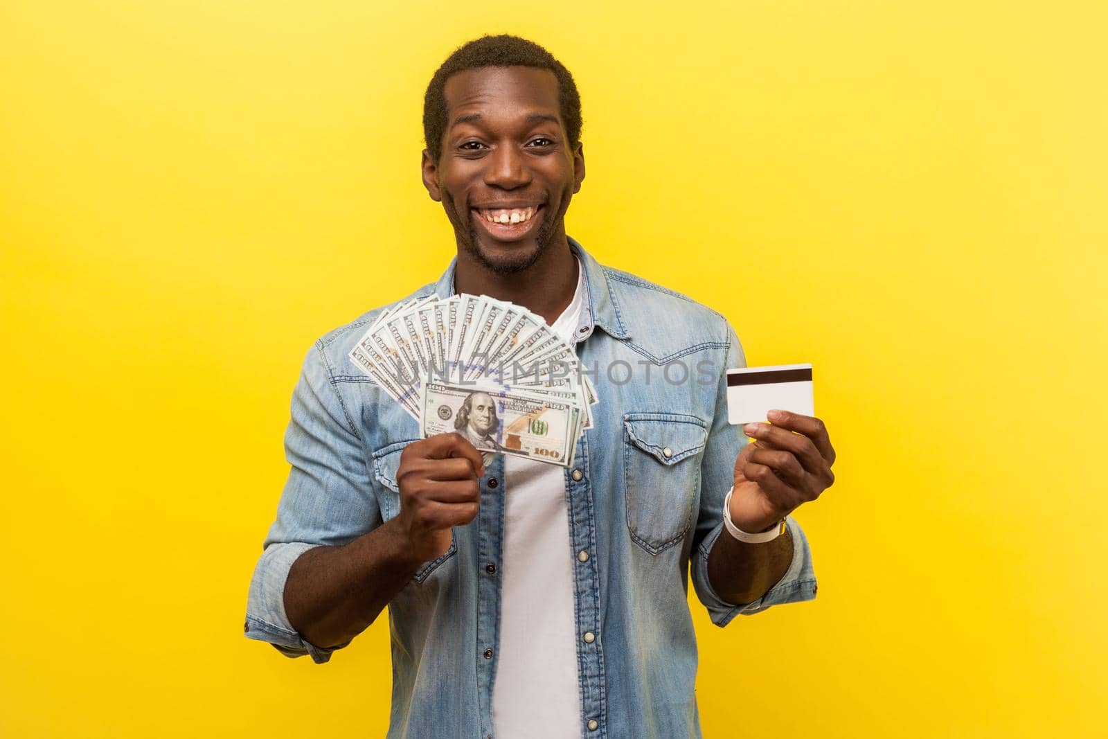 Portrait of excited young man in denim shirt holding dollar bills and credit card with satisfied expression, toothy smile, enjoying payments with cashback. studio shot isolated on yellow background