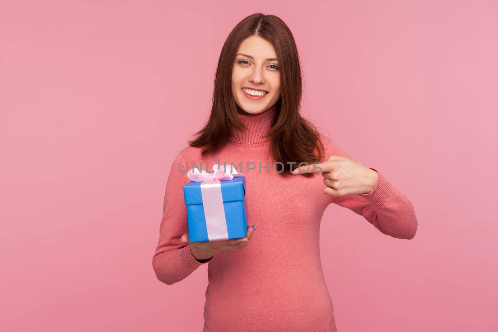 Satisfied happy woman with brown hair pointing finger on blue present with bow, looking at camera with toothy smile, suggesting bonuses. Indoor studio shot isolated on pink background
