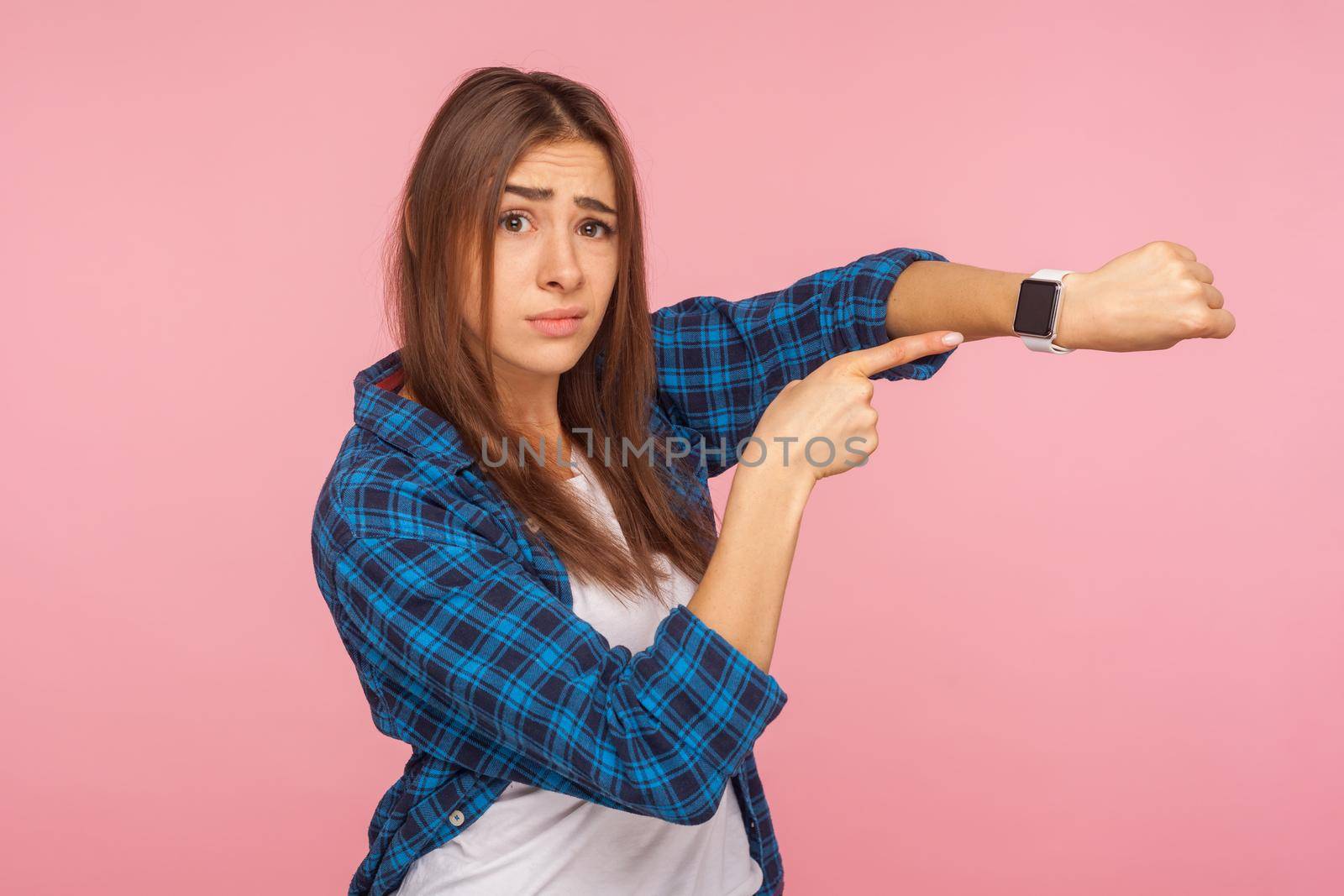 It's late. Portrait of impatient young woman in checkered shirt pointing at wrist watches checking time and looking worried, dissatisfied with delay. indoor studio shot isolated on pink background