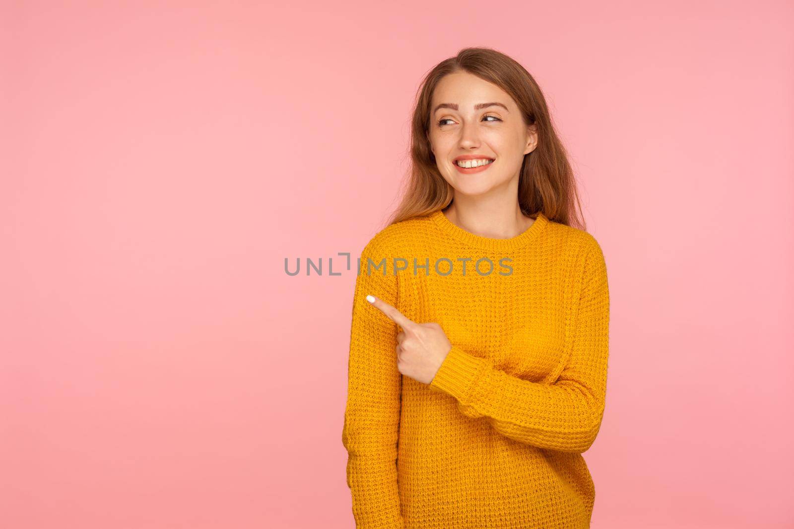 Look, attention. Portrait of cheerful attractive ginger girl in sweater pointing to the side and smiling, showing copy space, empty place for promotional text. studio shot isolated on pink background