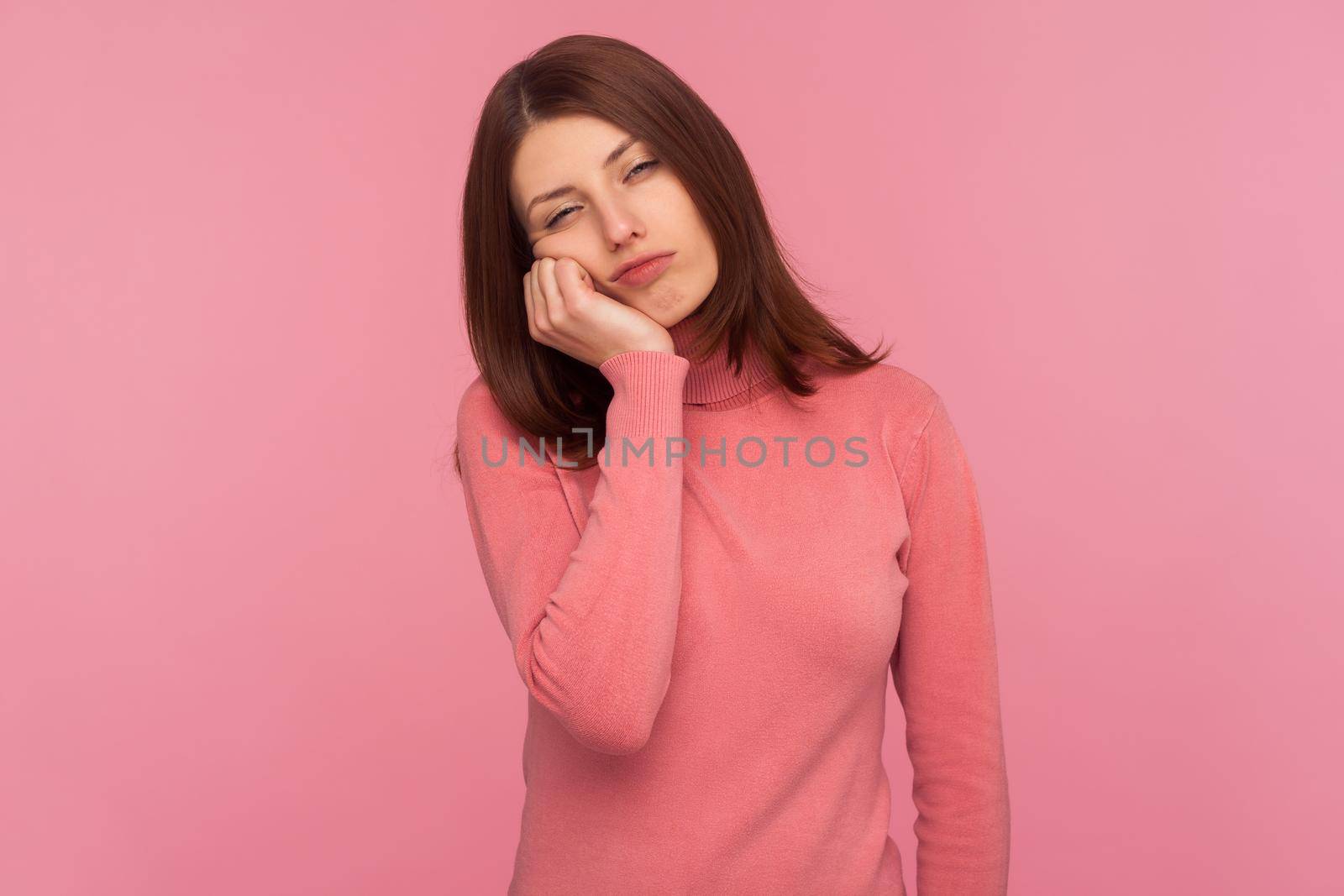 Lonely tired woman in pink sweater leaning head on hand, looking with bored expression, exhausted with overwork. Indoor studio shot isolated on pink background