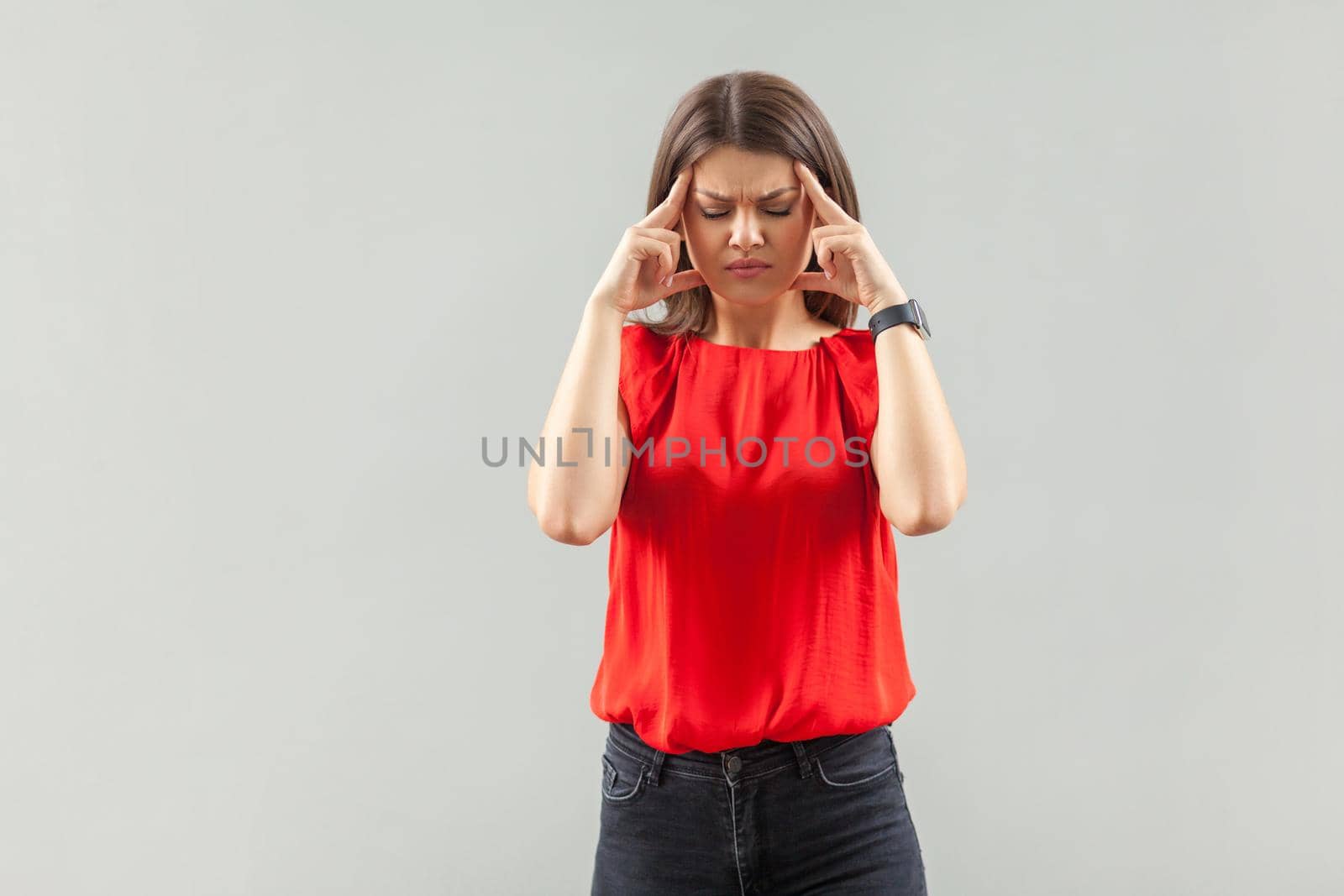 Headache, confution or thinking. Portrait of serious beautiful brunette young woman in red shirt standing and holding her painful head. indoor, studio shot, isolated on gray background.