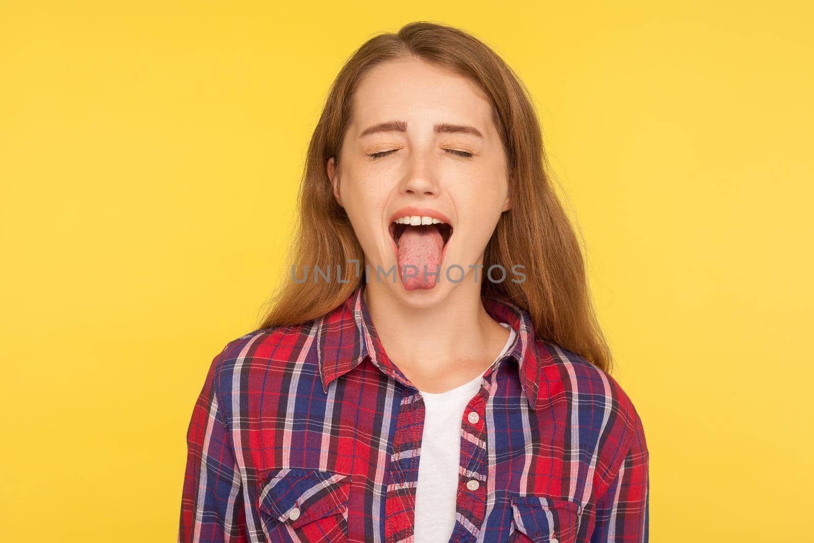 Portrait of naughty disobedient ginger girl in casual shirt standing with closed eyes and sticking out tongue, looking funny childish with derisive grimace. studio shot isolated on yellow background