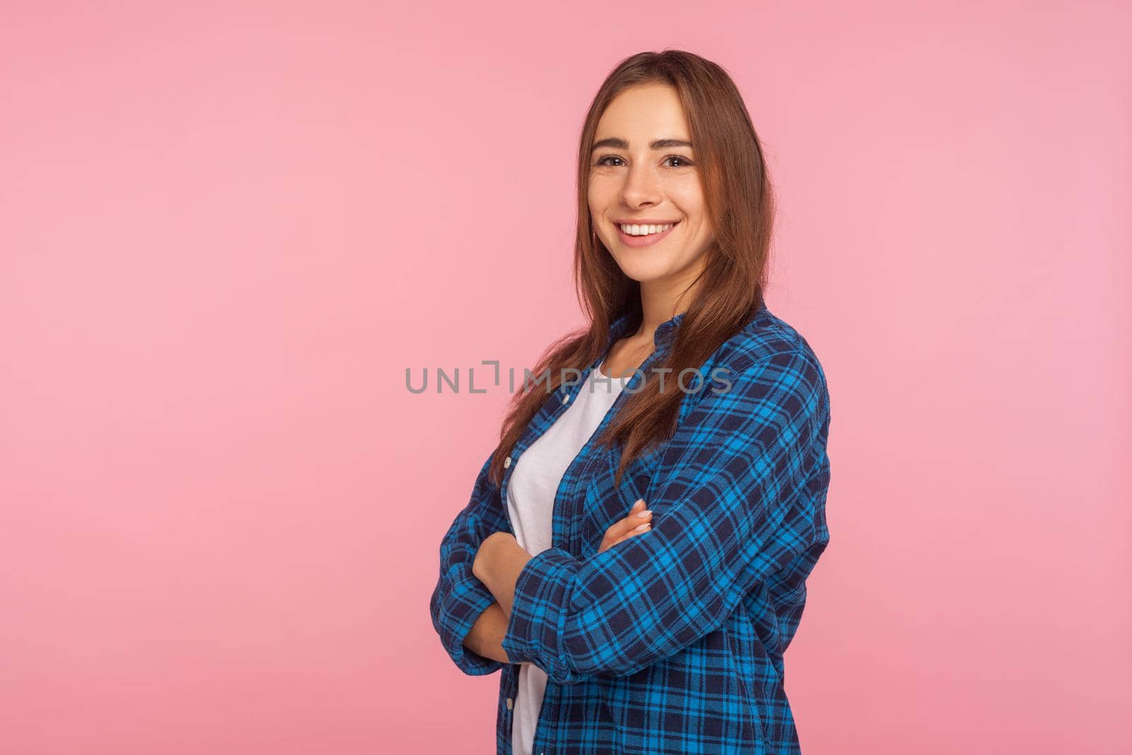 Portrait of cheerful girl in checkered shirt smiling sincerely at camera, holding hands crossed, looking confident and contented with life, rejoicing positive news. indoor studio shot, pink background