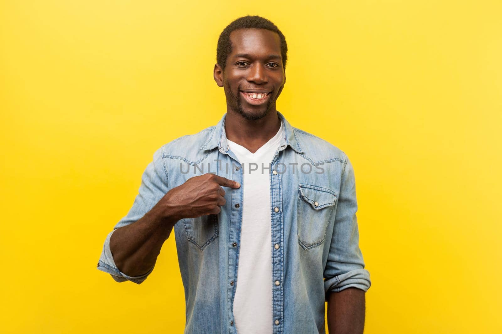 This is me. Portrait of successful positive young man in denim casual shirt pointing at himself, grinning happily at camera, rejoicing in achievement. indoor studio shot isolated on yellow background