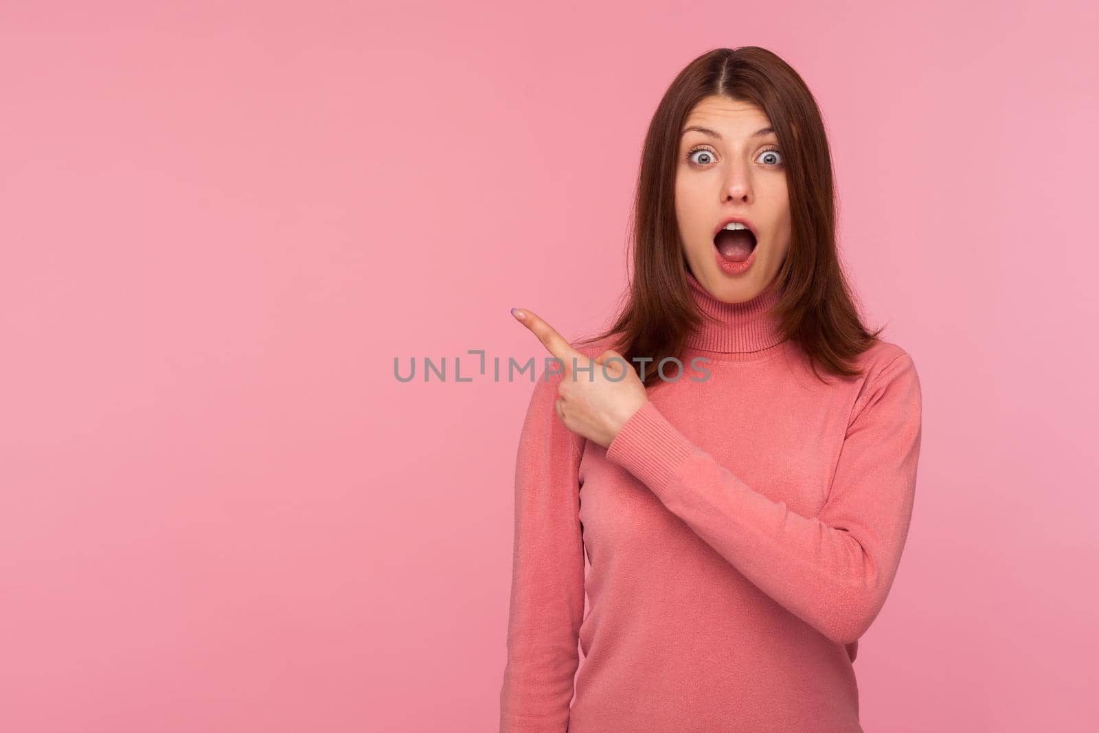 Extremely shocked brunette woman in pink sweater pointing away looking at camera with big eyes and open mouth, surprised with advertising, empty space. Indoor studio shot isolated on pink background
