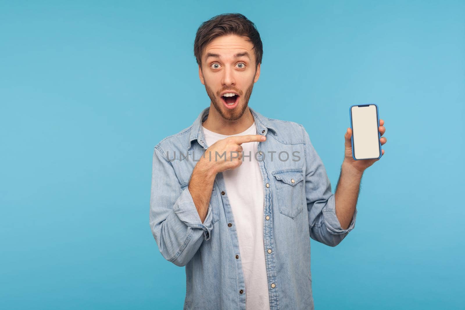 Portrait of amazed man standing with open mouth, shocked expression and showing mobile device, cell phone with mock up empty display to advertise. indoor studio shot isolated on blue background