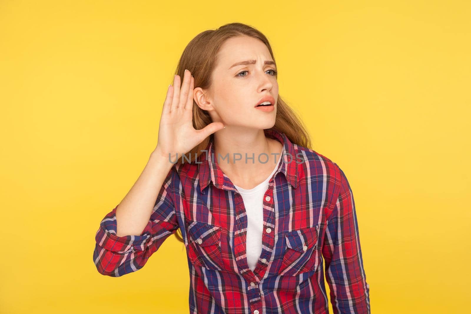 Can't hear you. Portrait of attentive ginger girl in checkered shirt holding hand near ear trying to listen quiet conversation, overhearing gossip. indoor studio shot isolated on yellow background