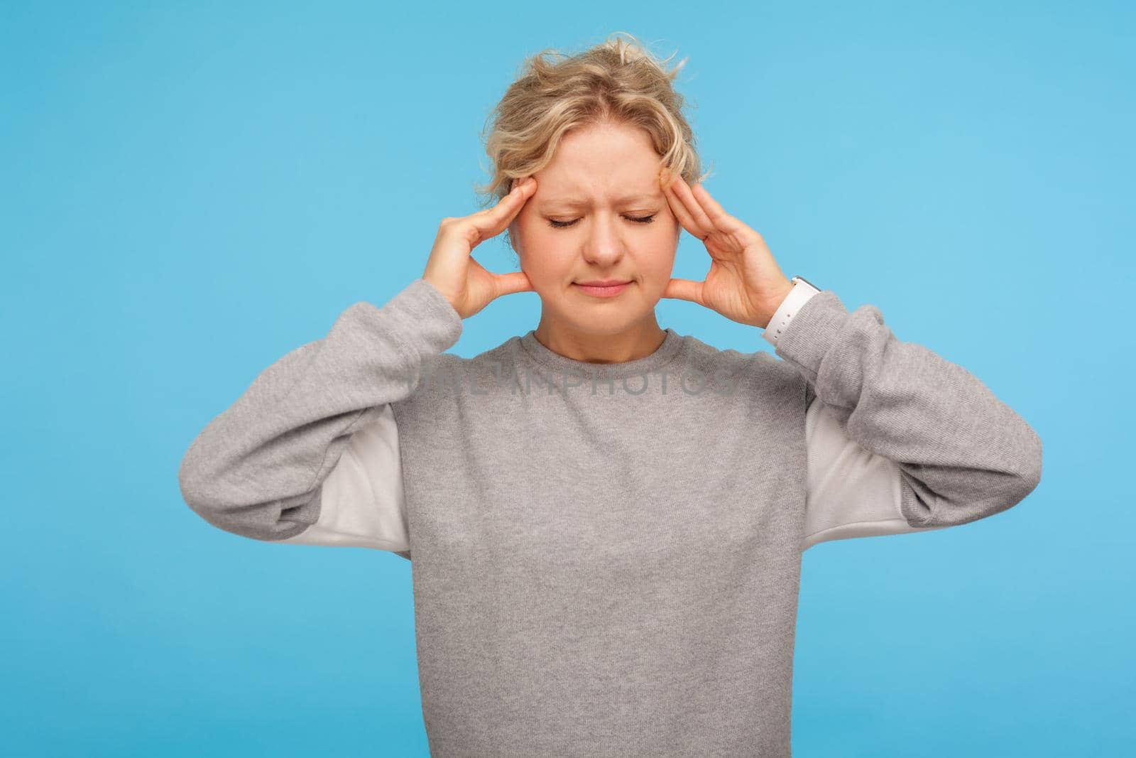 Portrait of depressed sick adult woman with short curly hair in sweatshirt holding head with hands, suffering headache, intense migraine of overwork. indoor studio shot isolated on blue background