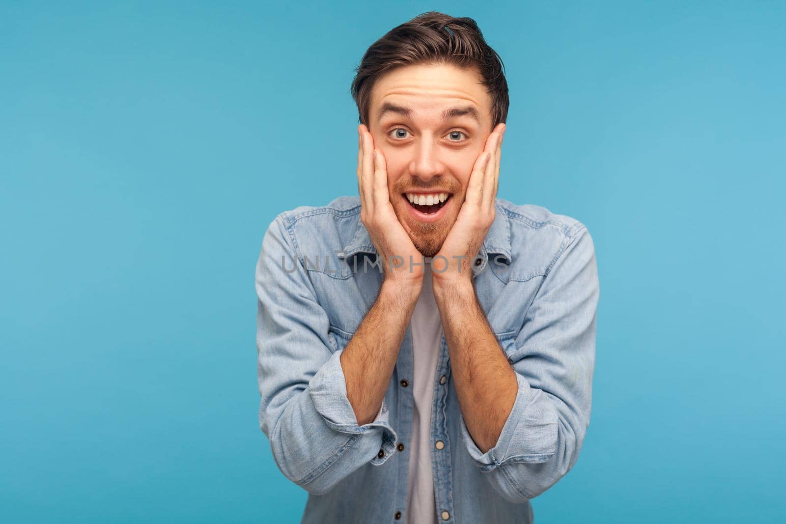 Wow, unbelievable. Astonished man in worker denim shirt grasping face in amazement, looking with open mouth, big eyes, feeling shocked and happy about unexpected success. indoor studio shot isolated