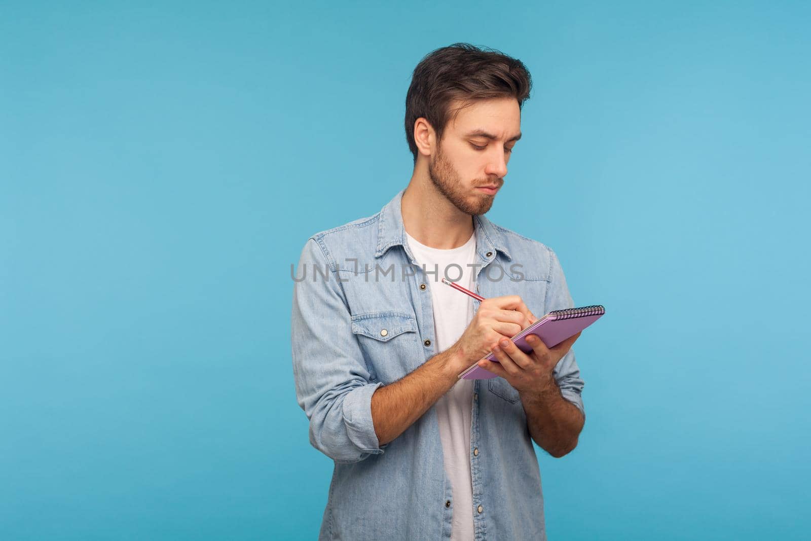 Portrait of man journalist in denim shirt making notes in paper notebook, writing business idea, future plans, checking appointment in schedule diary. indoor studio shot isolated on blue background