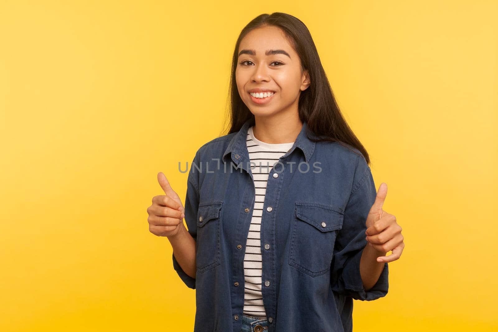Well done, awesome result. Portrait of happy girl in denim shirt showing thumbs up, gesturing like and smiling contented, satisfied with life, enjoying excellent service. indoor studio shot isolated