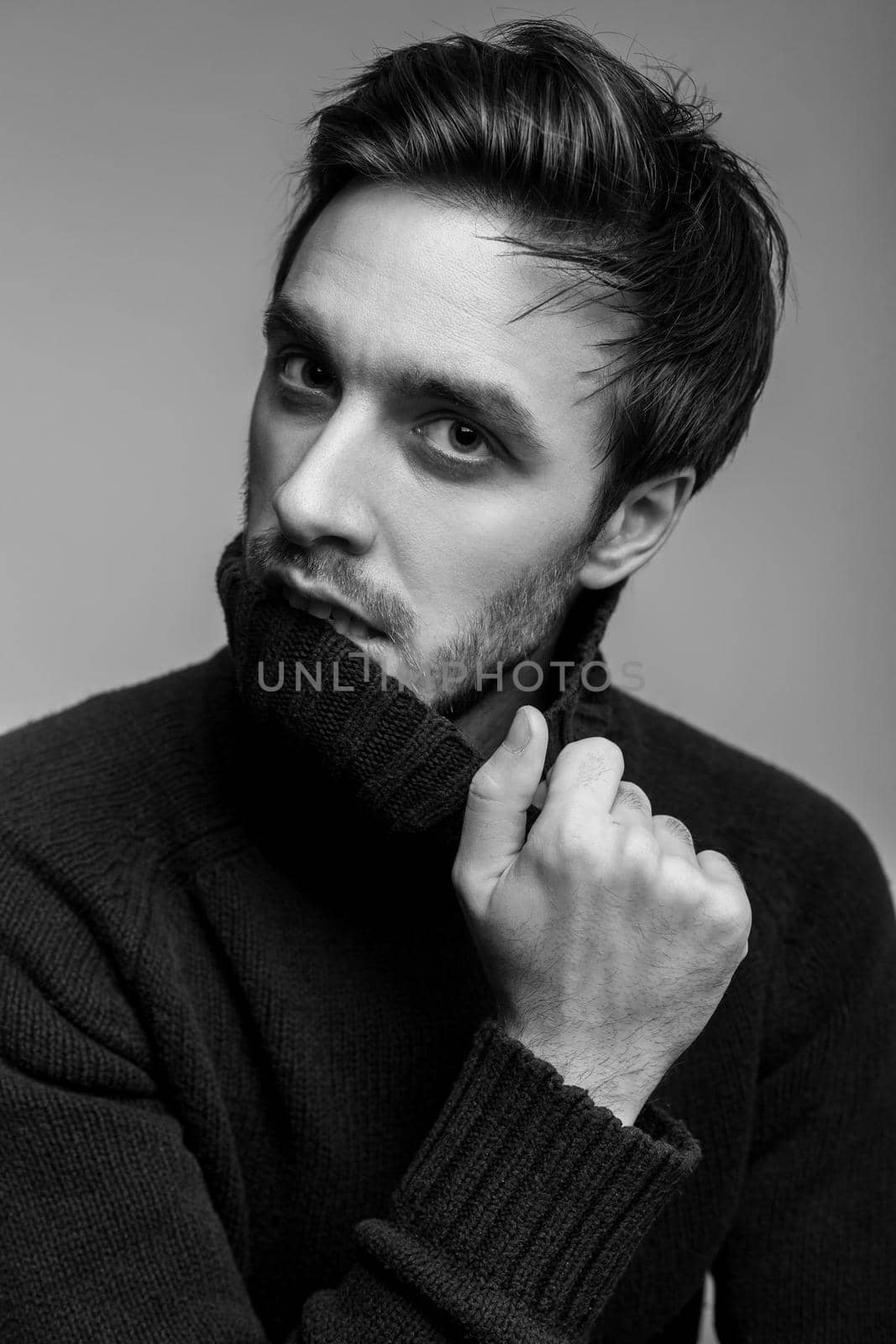 Portrait of sexy handsome man with stylish hairdo wearing warm sweater, biting high collar and looking seductive, flirting at camera, male fashion, vogue concept. black and white, studio shot isolated