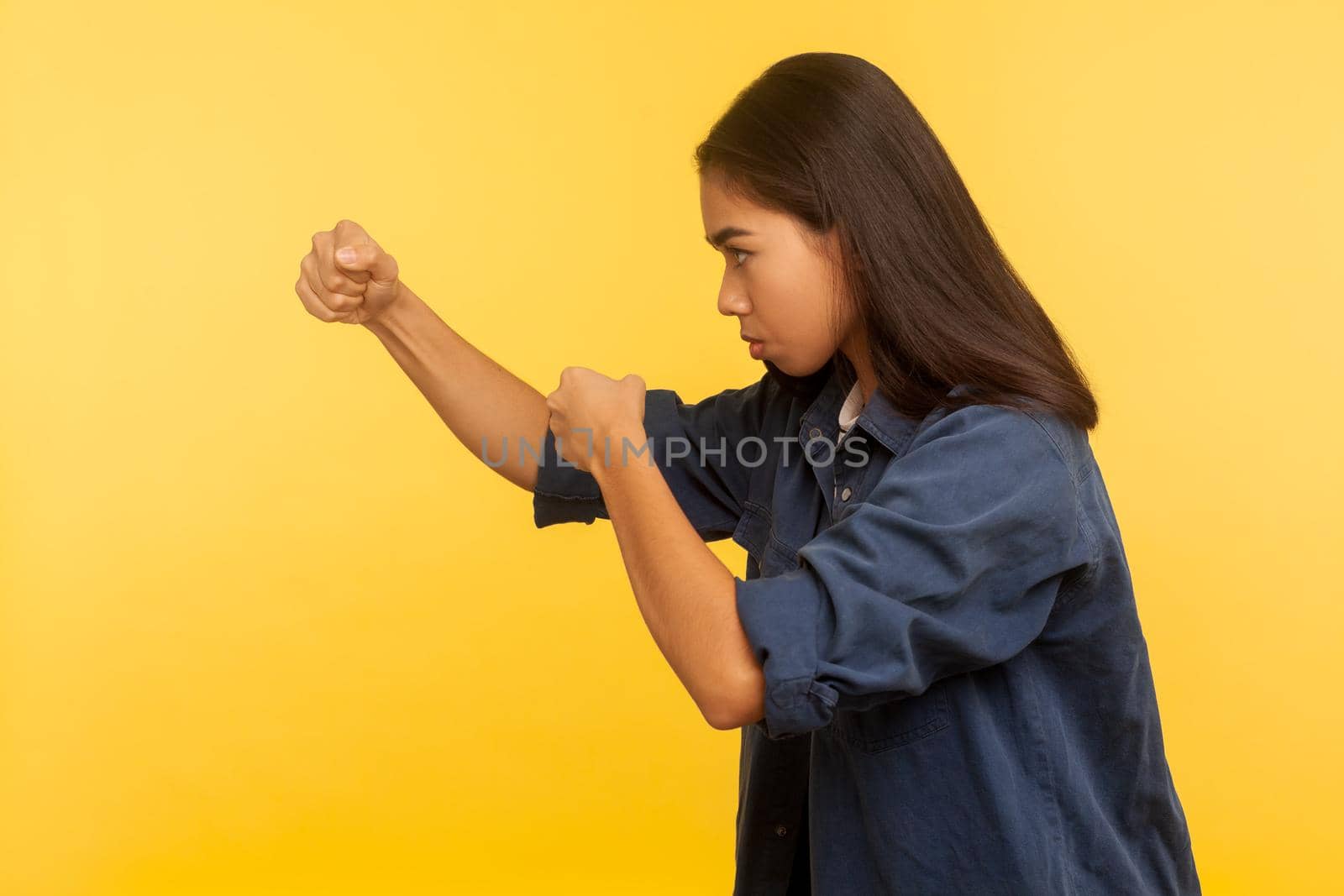 Let's fight. Side view of confident courageous girl in denim shirt keeping fists clenched, boxing and punching to side, struggle and self-defense concept. studio shot isolated on yellow background