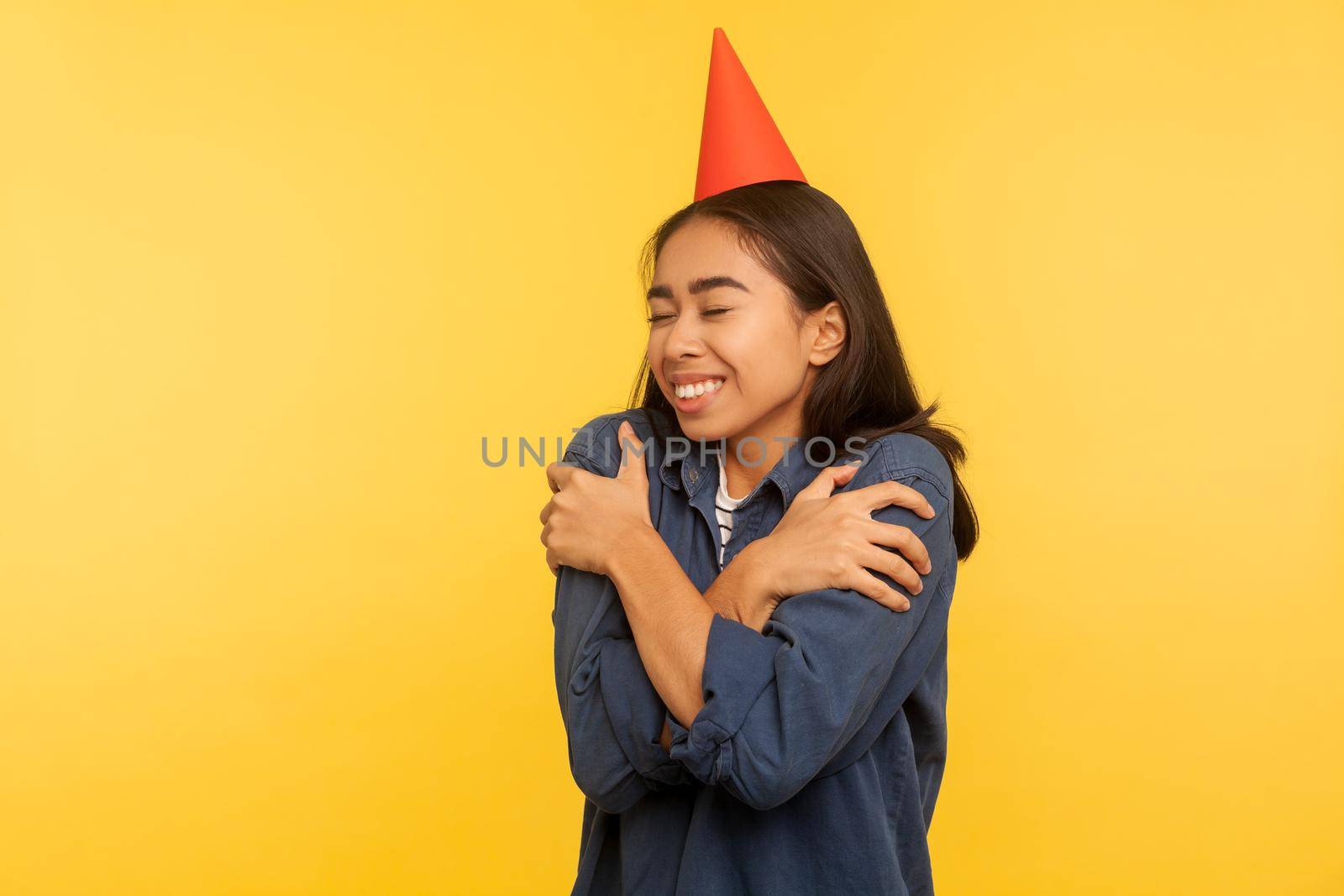 Happy birthday to me. Portrait of girl in denim shirt and with funny party cone on head embracing herself, celebrating successful event alone but happy. studio shot isolated on yellow background