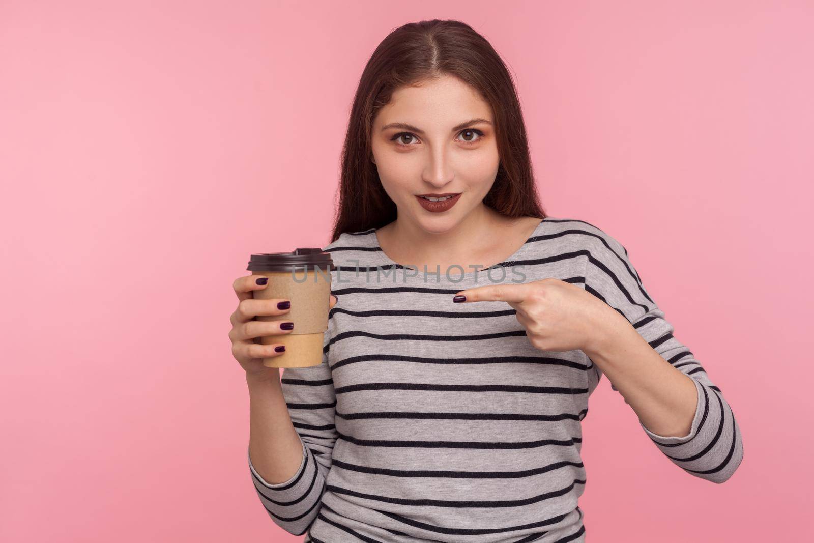 Portrait of attractive brunette woman in striped sweatshirt pointing at coffee and smiling to camera, drink with caffeine, energy boost in morning. indoor studio shot isolated on pink background