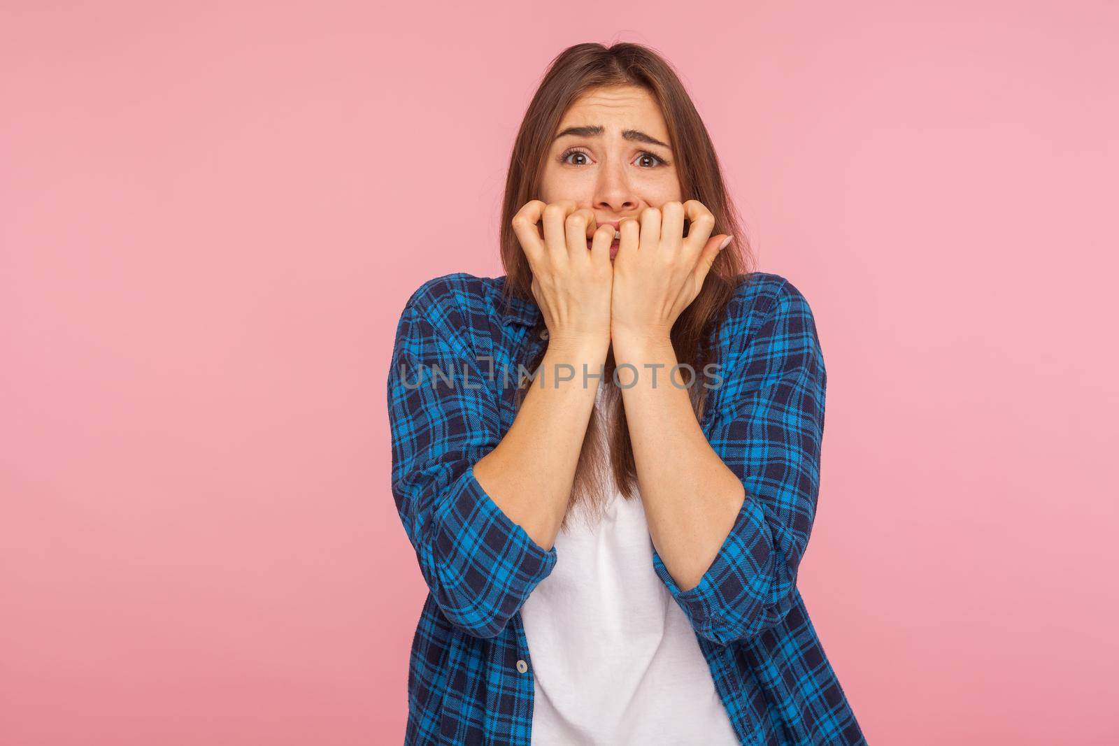 Anxiety. Portrait of scared girl in checkered shirt biting nails fingers and looking terrified, having depression and nervous problem, panic attack. indoor studio shot isolated on pink background