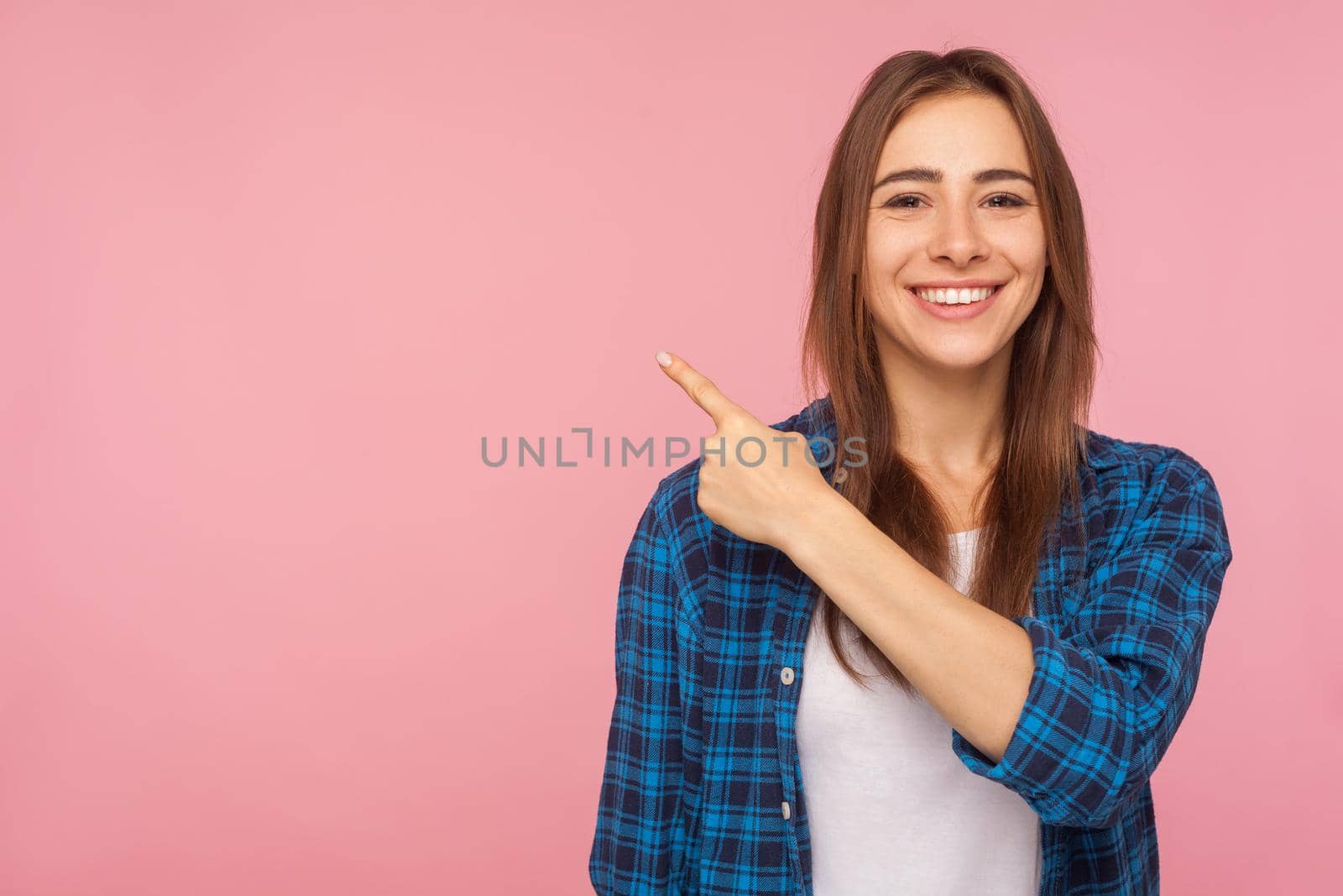 Look at advertisement. Portrait of charming brunette girl in checkered shirt pointing aside at empty place for ad content. studio shot isolated on pink background, copy space for commercial product