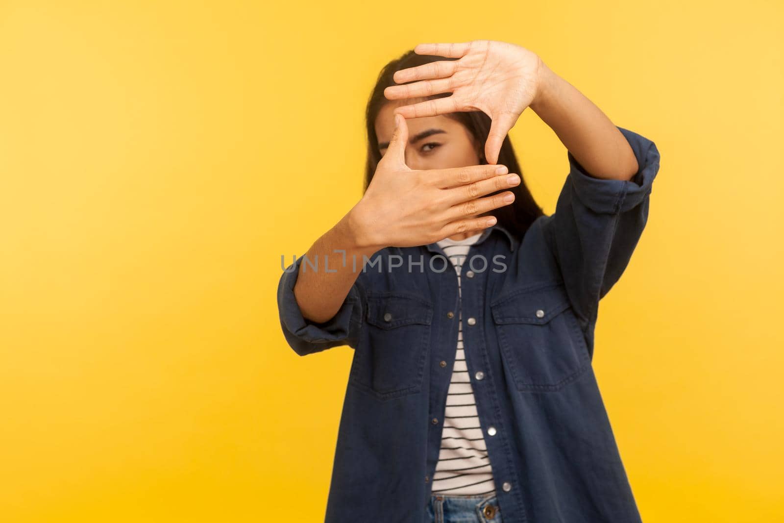 Portrait of girl in denim shirt looking through finger frame, imitating to take photo, focusing and cropping interesting moment, capturing moment. indoor studio shot isolated on yellow background