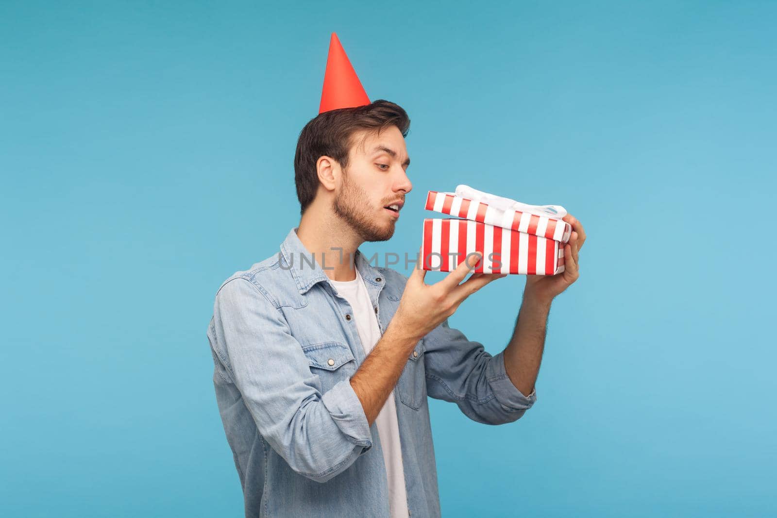 Portrait of curious man with party cone hat opening gift, peeking inside box with nosy look, unpacking present, in anticipation of interesting birthday surprise. studio shot isolated, blue background
