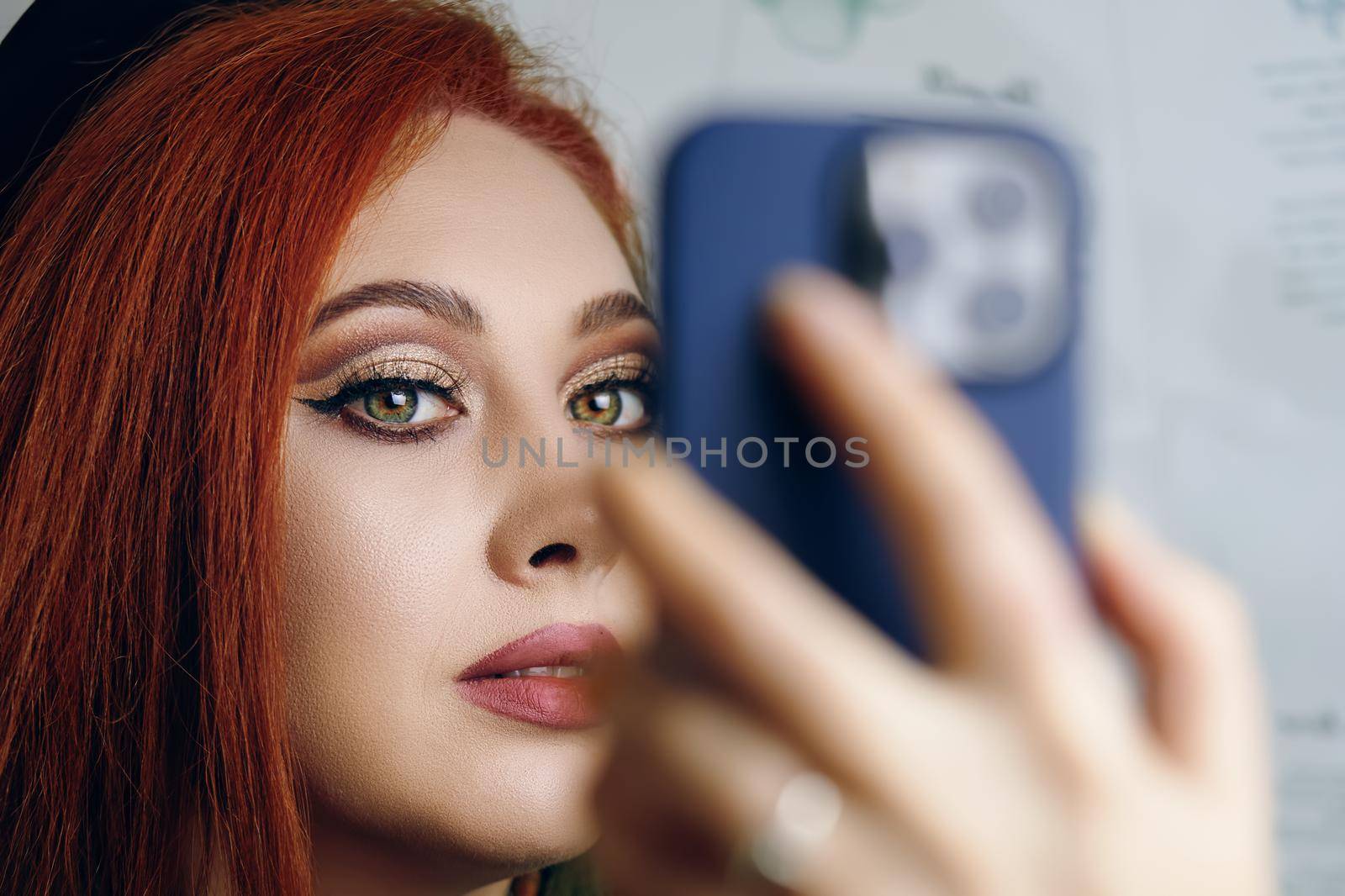 Red-haired woman with bright makeup takes a selfie on the camera of a modern phone. Blogger girl with a smartphone in her hand. Fashionable female hobby.