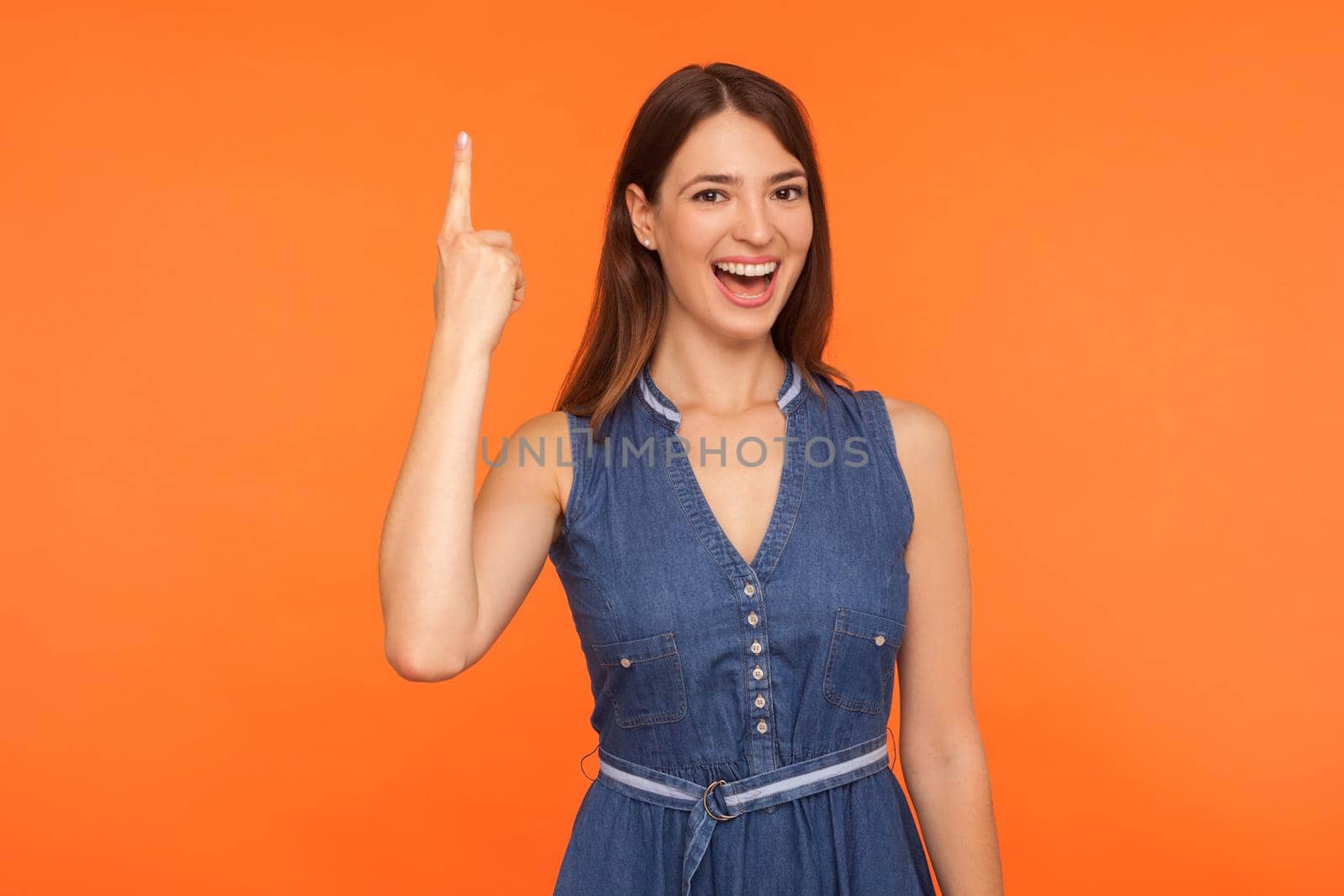 Smart brunette woman in denim dress pointing finger up with excellent idea, looking inspired and excited by sudden creative thought, found solution. indoor studio shot isolated on orange background