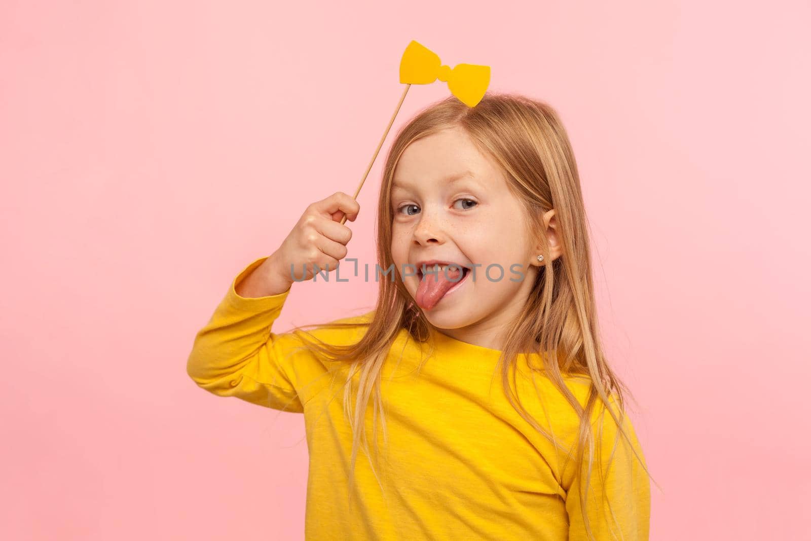 Portrait of playful adorable little ginger girl holding paper bow and sticking out tongue, naughty child having fun, wearing masquerade accessory. indoor studio shot isolated on pink background