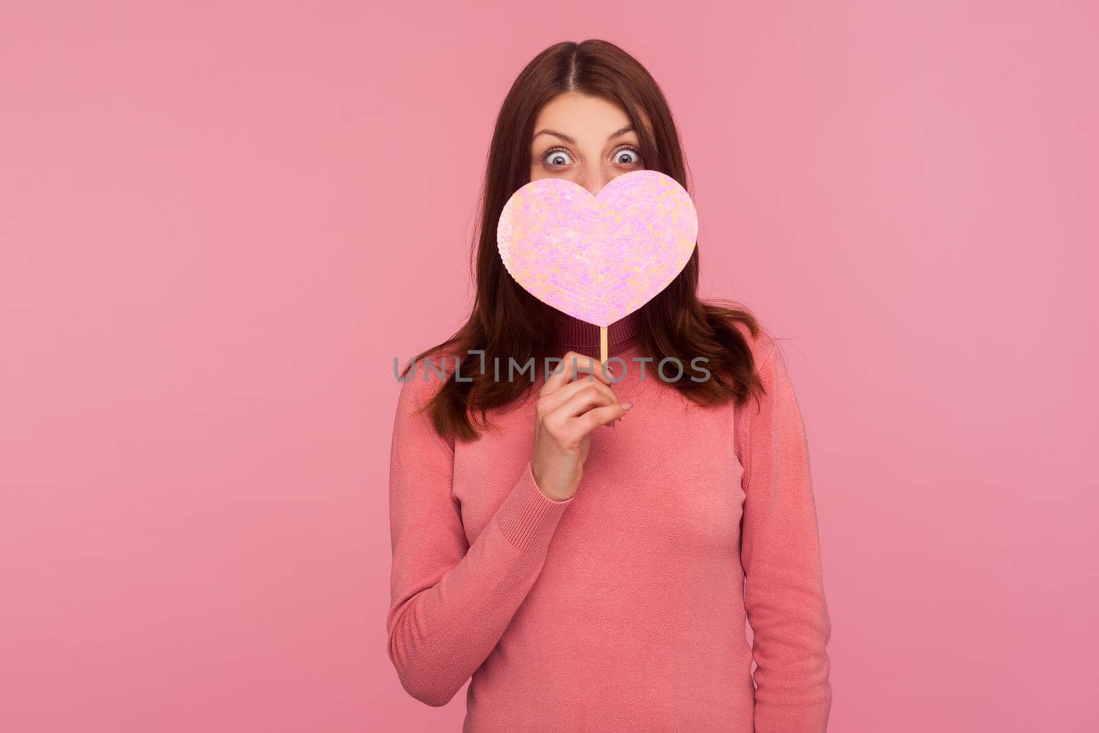 Surprised brunette woman in pink sweater covering face with pink heart, hiding behind symbol of love, wondered with romance date. Indoor studio shot isolated on pink background