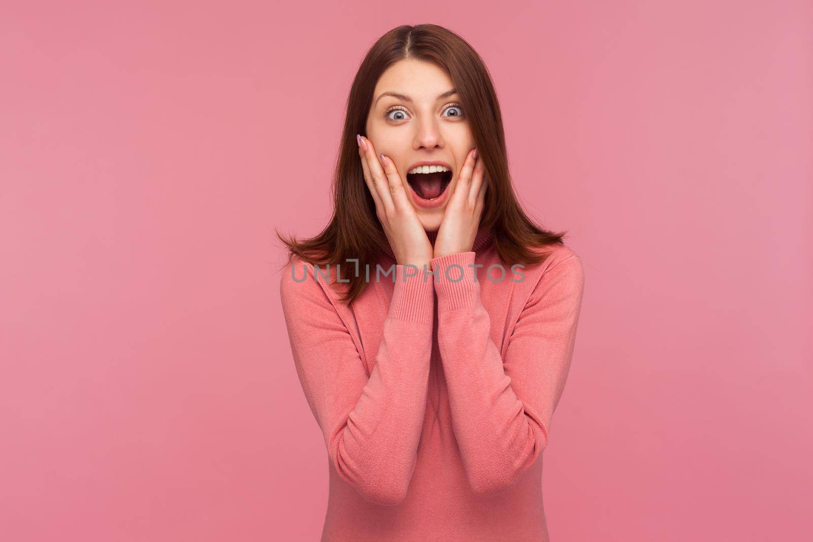 Extremely shocked surprised woman with brown hair screaming holding hands on cheeks, cant believe in her victory, astonishment. Indoor studio shot isolated on pink background