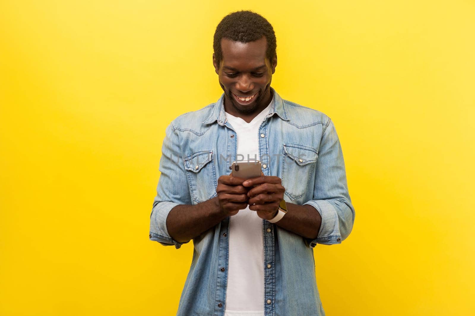 Portrait of happy smiley man in denim casual shirt using cellphone, typing text message or dialing number with joyous expression, technology concept. indoor studio shot isolated on yellow background