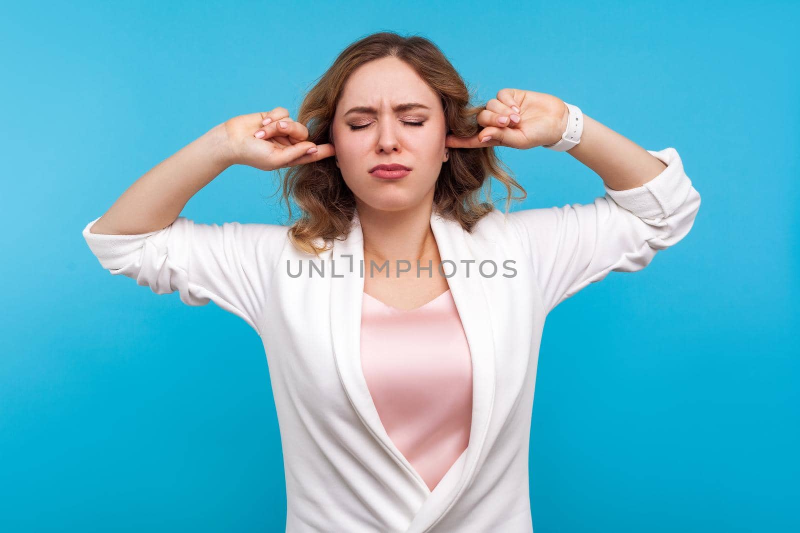 Don't want to listen, Portrait of resentful girl with wavy hair in white jacket standing covering ears and closed eyes in displeasure, frustrated about what she hear, ignoring loud noise. studio shot