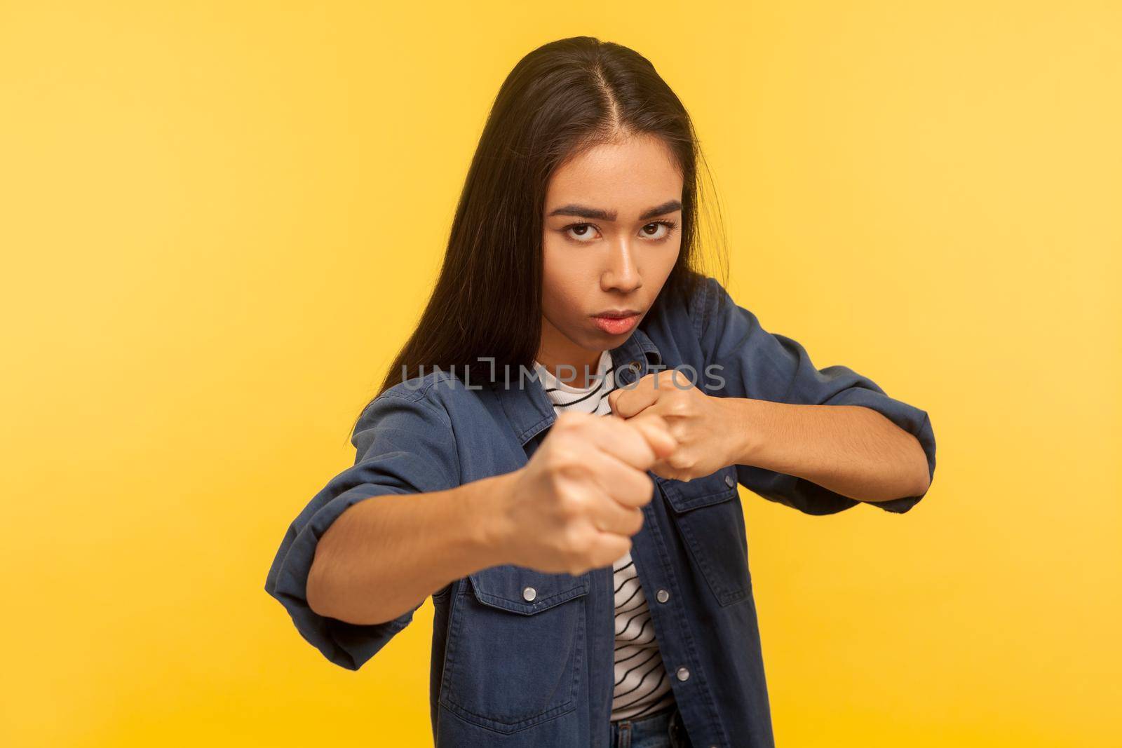 Let's fight. Portrait of confident courageous girl in denim shirt keeping fists clenched, boxing and punching at camera, her look expressing anger fury. studio shot isolated on yellow background