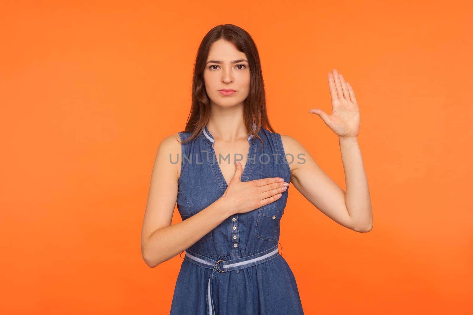 I swear. Dedicated honest brunette woman in denim dress standing with promise hand sign, gesturing palm up, giving promise, pledging allegiance. indoor studio shot isolated on orange background