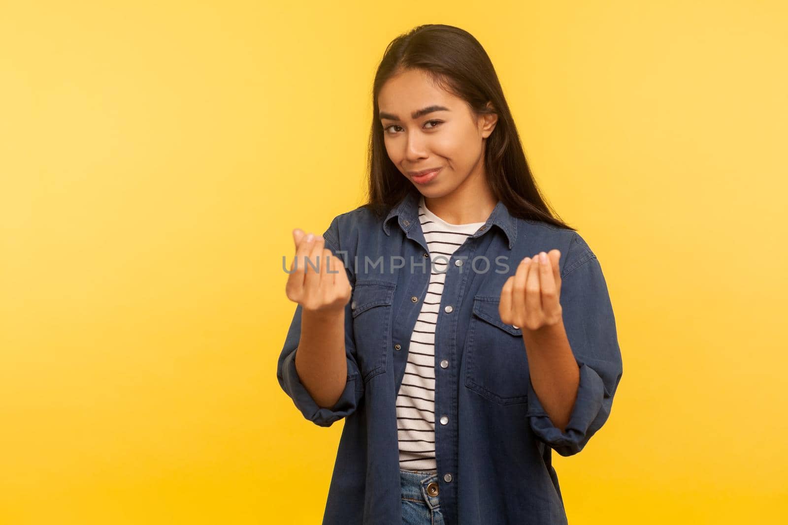 Portrait of greedy girl in denim shirt showing money gesture and looking with cunning smile, planning big profit, financial reward, demanding cash. indoor studio shot isolated on yellow background